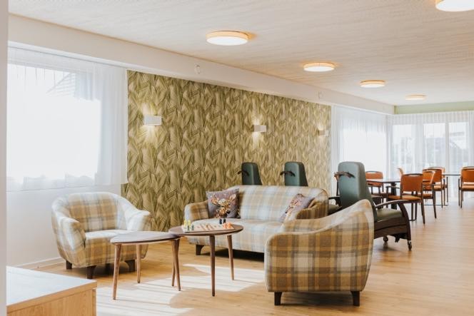 Gemeinschaftsraum der Tagespflege. Im Vordergrund Sitzgruppe mit Sesseln und im Hintergrund sind Ruhesessel und Tischgruppen zu sehen.